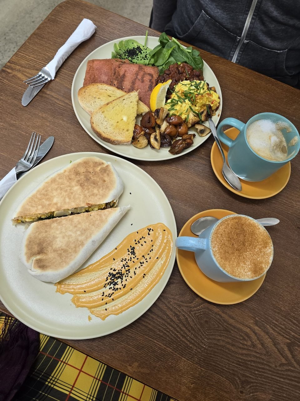 A big vegan breakfast spread with scramble, toast, plant-based 'facon' and greens, beside two cappuccinos.