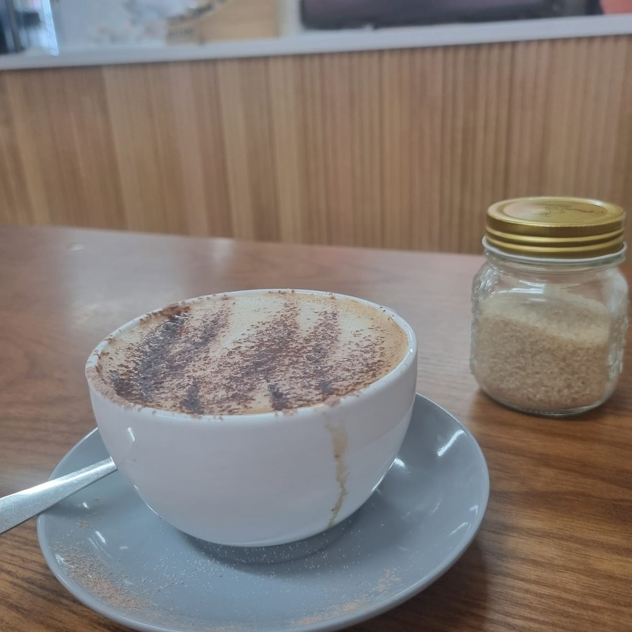 Cappuccino dusted with chocolate beside a jar of raw sugar against a timber wall.